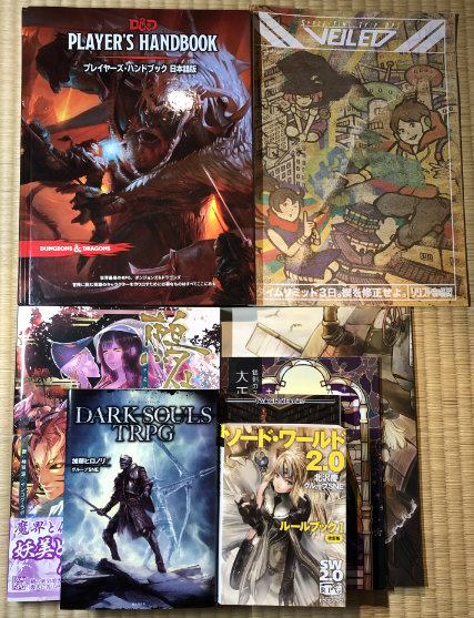 A variety of Japanese TRPGs stacked to show relative sizes. The only hardcover is the D&D 5e Player's Handbook.