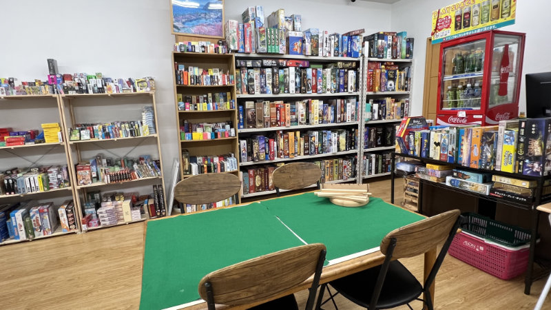 Table with green felt in the foreground and shelves of board games in the background. A drink fridge is to the right of the table.