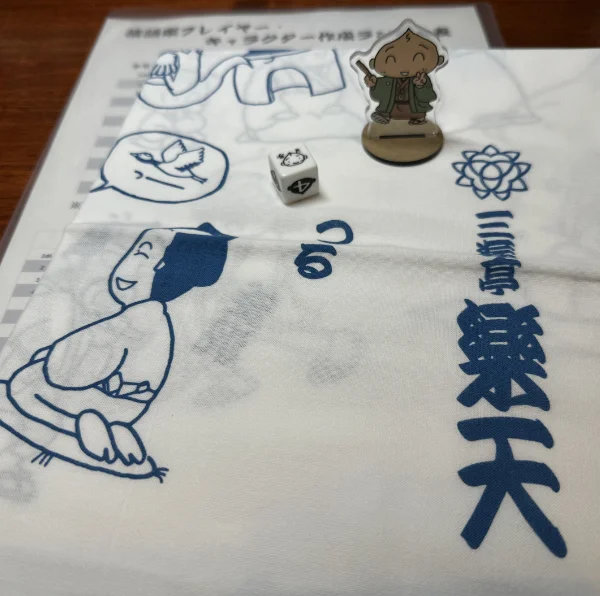 Goodies from the event. A figure with Rakuten's character is next to a die on top of a tenugui and paper sheet for generating rakugo characters.