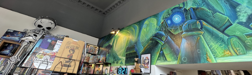 Ancient Robot Games - games zines on a rack and the counter in front of a large mural of a robot. Colors are mainly greens with blue highlights.