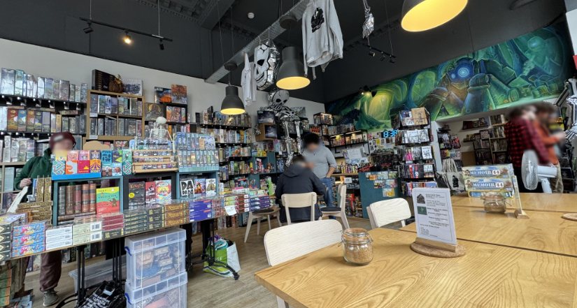 Photo of the store from a gaming table. Games for sale on the left and a game being played in the center. Store shirts are hanging above.