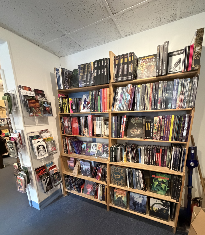 TTRPG books line two tall book cases. A smaller wall stand contains indie books.