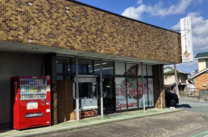Shopfront of Spinward Games. A large window graphic shows the logo. A vending machine is near the entrance.